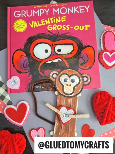 A children's book titled Grumpy Monkey: Valentine Gross-Out lies on a table surrounded by heart decorations, with a handmade Popsicle Stick Monkey puppet placed in front.