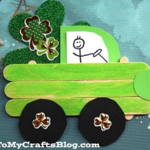 A Popsicle Stick St. Patrick's Day Truck craft features a green truck made from popsicle sticks, black wheels, glittery green shamrocks in the bed, and a stick figure driver—all set against a blue background with stars.