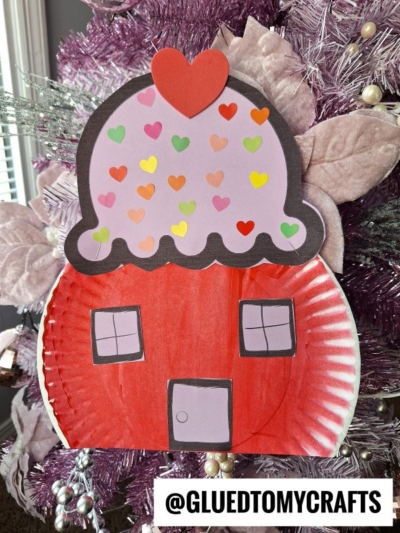 A paper plate cupcake house craft shaped like a red house with square windows, a purple roof adorned with colorful heart stickers, and a red heart on top. Displayed in front of a pink Christmas tree. Text reads @GLUEDTOMYCRAFTS.