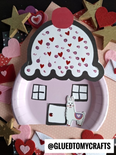 A whimsical paper plate cupcake house craft in pink, adorned with hearts, sequins, and a llama cutout. Cutout hearts and stars surround the piece. The text @GLUEDTOMYCRAFTS appears at the bottom.