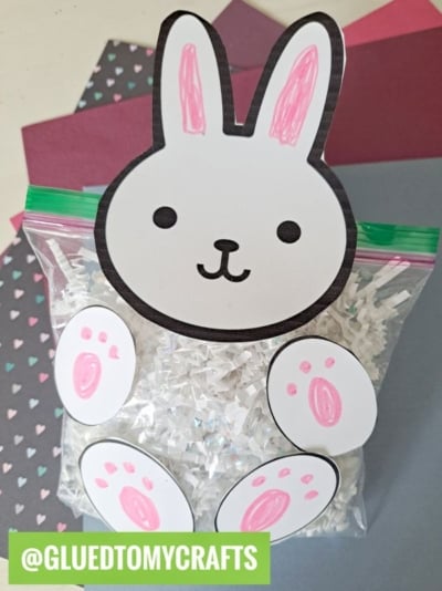 A clear plastic bag filled with crinkle paper bunny shreds is decorated with a cute bunny face and paws made of paper. Pink and purple craft paper with heart patterns are in the background. Text reads @GLUEDTOMYCRAFTS.
