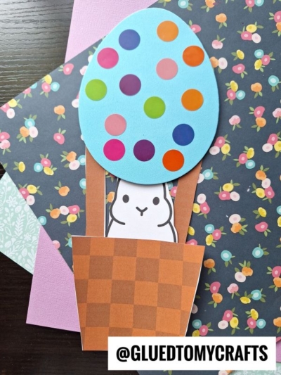 A paper craft featuring a white bunny in a brown basket with a large, blue, polka-dotted easter egg hot air balloon on floral and pastel papers. Text reads @GLUEDTOMYCRAFTS at the bottom.