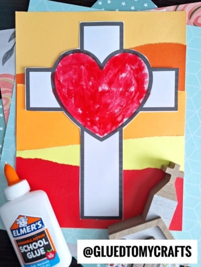 An art project featuring a white cross with a red heart in the center, set against a torn paper Easter sunrise of layered orange and yellow, with a bottle of glue, wooden cross, and @GLUEDTOMYCRAFTS at the bottom.