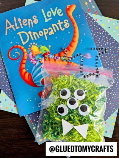 A children’s book titled Aliens Love Dinopants rests on patterned paper beside a clear bag filled with green crinkle paper crafts, five googly eyes, and white paper shapes forming a monster mouth. @GLUEDTOMYCRAFTS is labeled.