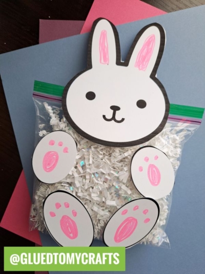 A craft project of a crinkle paper bunny made from a ziplock bag filled with white paper shreds. The bag has paper ears, face, and paws attached with pink details. The background features blue and pink paper. @GLUEDTOMYCRAFTS is labeled.