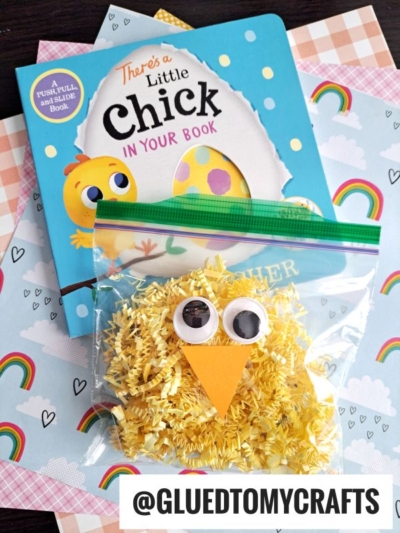 A children’s book titled “There’s a Little Chick in Your Book” sits on rainbow-themed paper next to a bag of yellow pasta crafted to look like a chick using crinkle paper crafts, with googly eyes, an orange beak, and the handle @GLUEDTOMYCRAFTS shown.