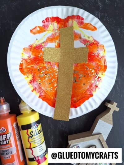 A paper plate decorated with orange and yellow paint splat easter sunrise is topped with a gold glitter cross. Bottles of orange and yellow paint and a small wooden cross are nearby. The text @GLUEDTOMYCRAFTS appears at the bottom.