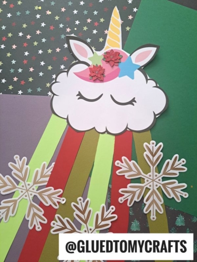 A rainbow unicorn cloud craft made of paper features closed eyes, a horn, pink flowers, and colorful streamers. Surrounded by paper snowflakes on patterned backgrounds, it’s a magical DIY. Text at the bottom reads @GLUEDTOMYCRAFTS.