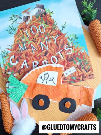 A children’s book titled Too Many Carrots is displayed with a handmade orange Paper Plate Bunny Car, fake carrots, a bunny tail, and bunny ear decorations surrounding it. The Instagram handle @GLUEDTOMYCRAFTS appears at the bottom.