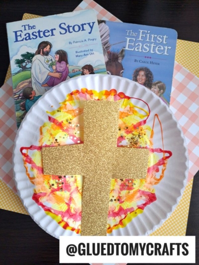 A paper plate with colorful paint, resembling a marbled sunrise craft and a large gold glitter cross, is displayed in front of two Easter-themed children’s books titled The Easter Story and The First Easter. A yellow checked paper sits beneath the plate.