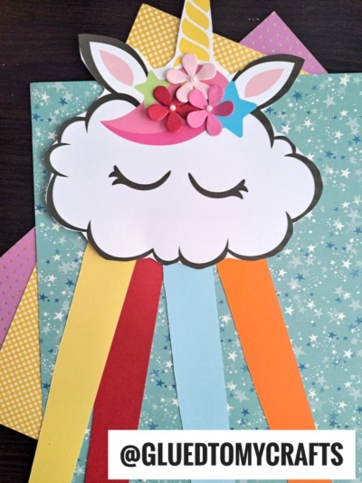 A whimsical rainbow unicorn cloud craft featuring a cloud face with closed eyes, colorful paper flowers, a yellow horn, and rainbow paper strips on layered patterned papers. The handle @GLUEDTOMYCRAFTS is visible.