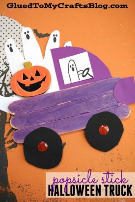 A Popsicle Stick Halloween Truck made of purple popsicle sticks with black wheels, carrying a smiling pumpkin and popsicle stick ghost cutouts in the back, sits on an orange background with a haunted tree design.