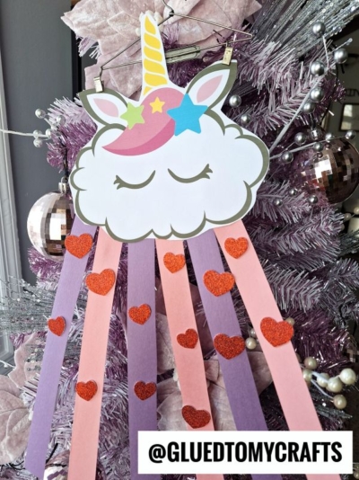 A rainbow unicorn cloud with a horn, ears, and closed eyes is decorated with pink and purple streamers and red glitter hearts, hanging on a lavender Christmas tree. Text reads @GLUEDTOMYCRAFTS.
