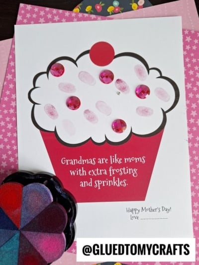 A handmade Mother’s Day card features a cupcake with pink jewel “sprinkles,” the phrase “grandmas are like moms with extra frosting and sprinkles,” and space to sign. A stamp pad and pink paper complete this sweet tribute.