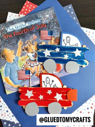 Two popsicle stick parade float crafts—one red, one blue with stars—sit on blue paper by a patriotic children’s book, mini US flags, and star-patterned paper. Each float looks like a popsicle stick patriotic truck with a stick-figure drawing inside.