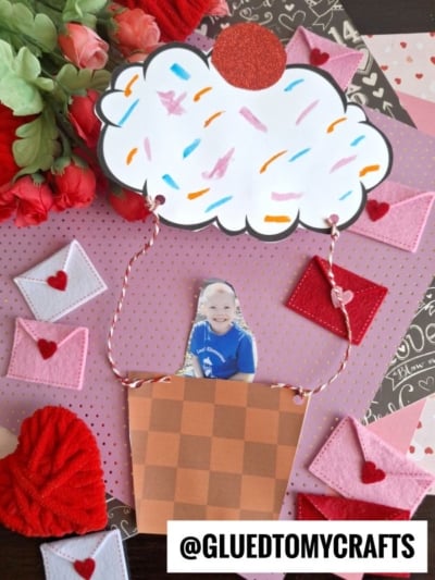 A handmade Valentine’s craft features a paper cupcake hot air balloon with a basket holding a child’s photo, surrounded by hearts, flowers, and mini felt envelopes. The handle reads @GLUEDTOMYCRAFTS.