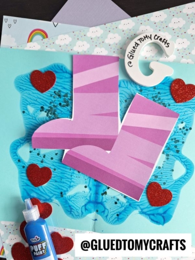 Colorful craft project with two pink-striped paper rain boots, a playful paint splat rain puddle, red glitter hearts, blue puffy paint, and a round Glued To My Crafts tag on a light blue background. @GLUEDTOMYCRAFTS text is at the bottom.