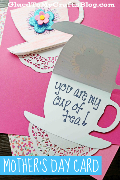 A handmade Mother’s Day paper teacup card sits on pink paper. Open, it reveals the handwritten message, You are my cup of tea! A flower and doily decorate the charming teacup.