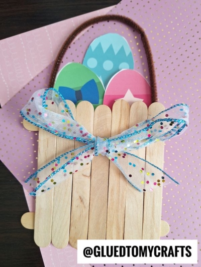 A handmade popsicle stick Easter egg basket features a colorful bow and a pipe cleaner handle, holding three paper eggs. Placed on pink patterned paper, the text @GLUEDTOMYCRAFTS appears at the bottom.