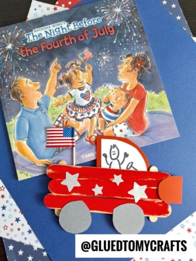 A children’s book titled “The Night Before the Fourth of July” is shown with a handmade popsicle stick patriotic truck craft featuring silver stars and paper wheels, set on blue paper with patriotic star-patterned paper nearby.