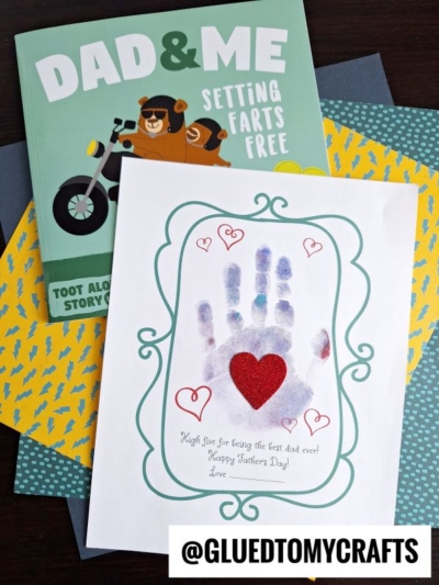 A handmade Father’s Day card featuring a child’s handprint high five and a red heart sits on patterned paper beside the funny book “Dad & Me: Setting Farts Free.” The Instagram handle @GLUEDTOMYCRAFTS is also visible.