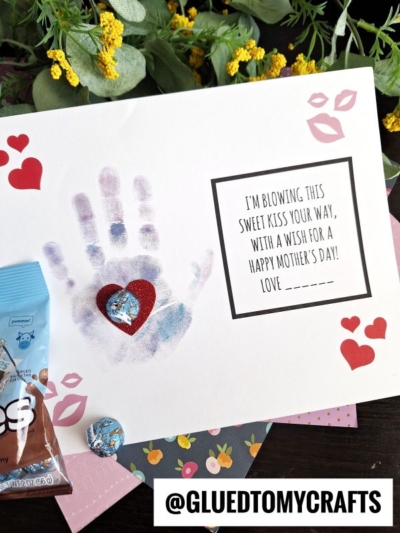 A handmade Mother’s Day card with a child’s handprint, heart stickers, a chocolate heart, lip print stickers, and a note saying, “I’m blowing kisses for mommy your way, with a wish for a Happy Mother’s Day! Love _____.”.