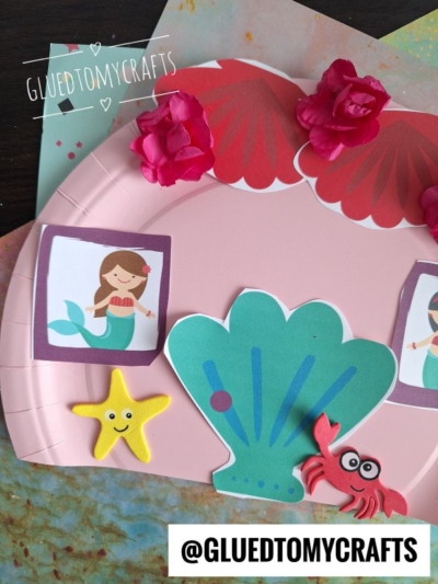 A pink paper plate mermaid house craft decorated with paper flowers, a shell, a starfish, a crab, and mermaid illustrations. The background features colorful paper, with @GLUEDTOMYCRAFTS written at the bottom.