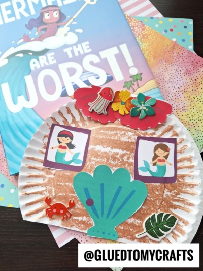 A brown, textured paper plate mermaid house craft decorated with cutouts of mermaids, a blue shell, a crab, a jellyfish, flowers, and leaves. Colorful papers and the book Mermaids Are The Worst! are in the background.