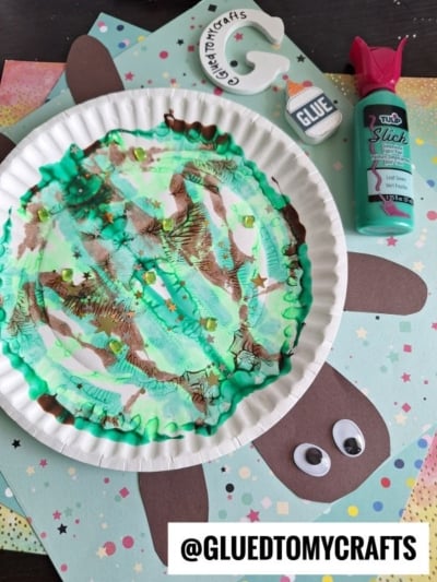 A paper plate turtle decorated with green, brown, and teal paint sits on colorful craft paper. Also visible are a bottle of teal paint, a glue sticker, googly eyes, and a bunny face made from brown and white paper.