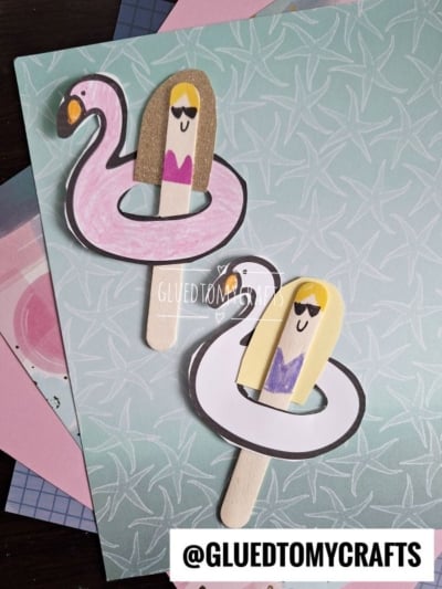 Two craft stick figures with paper bodies sit in drawn popsicle stick pool floats—one pink flamingo and one white swan—each with a smiling person wearing sunglasses, on patterned pastel paper backgrounds. A fun pool float craft for kids!.