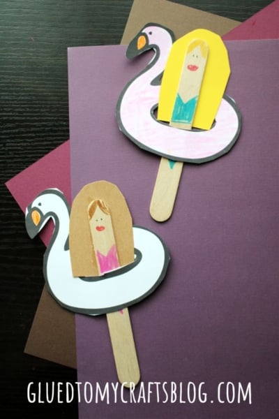 Two craft stick figures with paper bodies sit in drawn popsicle stick pool floats—one pink flamingo and one white swan—each with a smiling person wearing sunglasses, on patterned pastel paper backgrounds. A fun pool float craft for kids!.