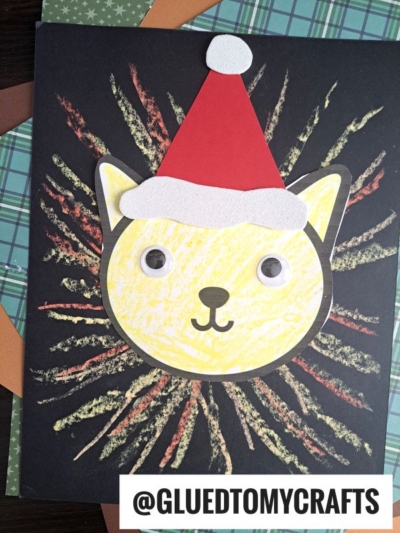 A handmade Christmas card features a Chalk Art Lion—a yellow cat face with googly eyes and a red Santa hat, surrounded by colorful streaks on a black background. The username @GLUEDTOMYCRAFTS is at the bottom.