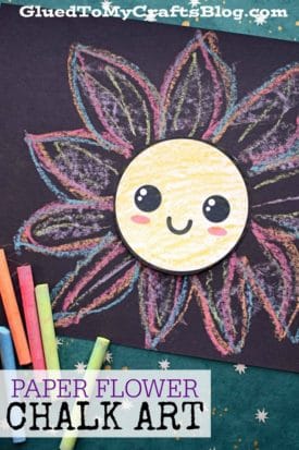 A cheerful chalk art flower with a smiling face sits on black paper, surrounded by colorful pastel chalk petals. Pieces of chalk lie nearby. Text reads, PAPER FLOWER CHALK ART.