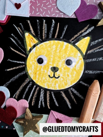 A yellow cat face drawing with googly eyes and a black outline is surrounded by radiating lines, evoking a Chalk Art Lion. Felt hearts, stars, envelopes, and pastel crayons decorate the edges. Text reads @GLUEDTOMYCRAFTS at the bottom.