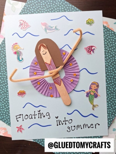 A handmade card shows a popsicle stick girl pool lounging with a purple cupcake liner float and tan pipe-cleaner arms, surrounded by sea-themed stickers and wavy lines. The text reads “Floating into summer.”.
