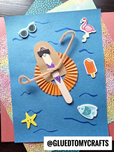 A popsicle stick craft of a girl with brown hair and sunglasses lounges on an orange cupcake liner pool float, surrounded by summer-themed stickers—perfect for making with popsicle stick pool lounging friends.