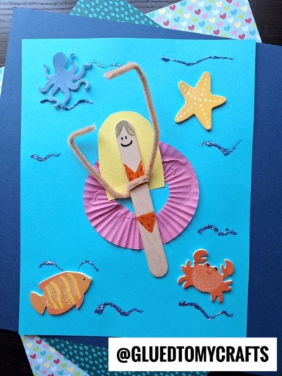A handmade mermaid craft featuring popsicle stick pool lounging friends and paper decorations of an octopus, starfish, fish, and crab on a blue background with wavy glitter lines. @GLUEDTOMYCRAFTS.