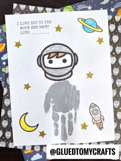 A handmade space-themed card features a Handprint Astronaut Keepsake with a gray handprint body, surrounded by stars, a planet, rocket, and moon. Text at the top reads, I love you to the moon and back! Love ____.