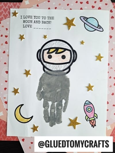 A handmade card features a Handprint Astronaut Keepsake, surrounded by cut-out stars, a moon, rocket, and planet. Text reads, I LOVE YOU TO THE MOON AND BACK! LOVE ____. @GLUEDTOMYCRAFTS is at the bottom.