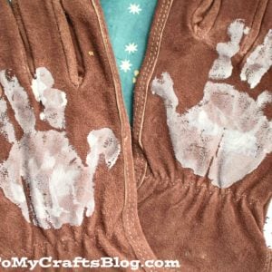 A pair of brown handprint work gloves with white handprints painted on the palms, displayed on a blue surface. The gloves are labeled GluedToMyCraftsBlog.com at the bottom.