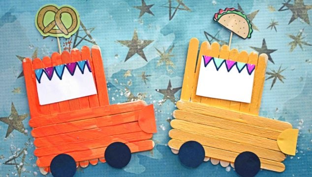 Two colorful popsicle stick food trucks made from craft sticks and paper are displayed on a blue starry background—one orange with a pretzel, the other yellow with a taco. Both feature black wheels and festive paper banners.