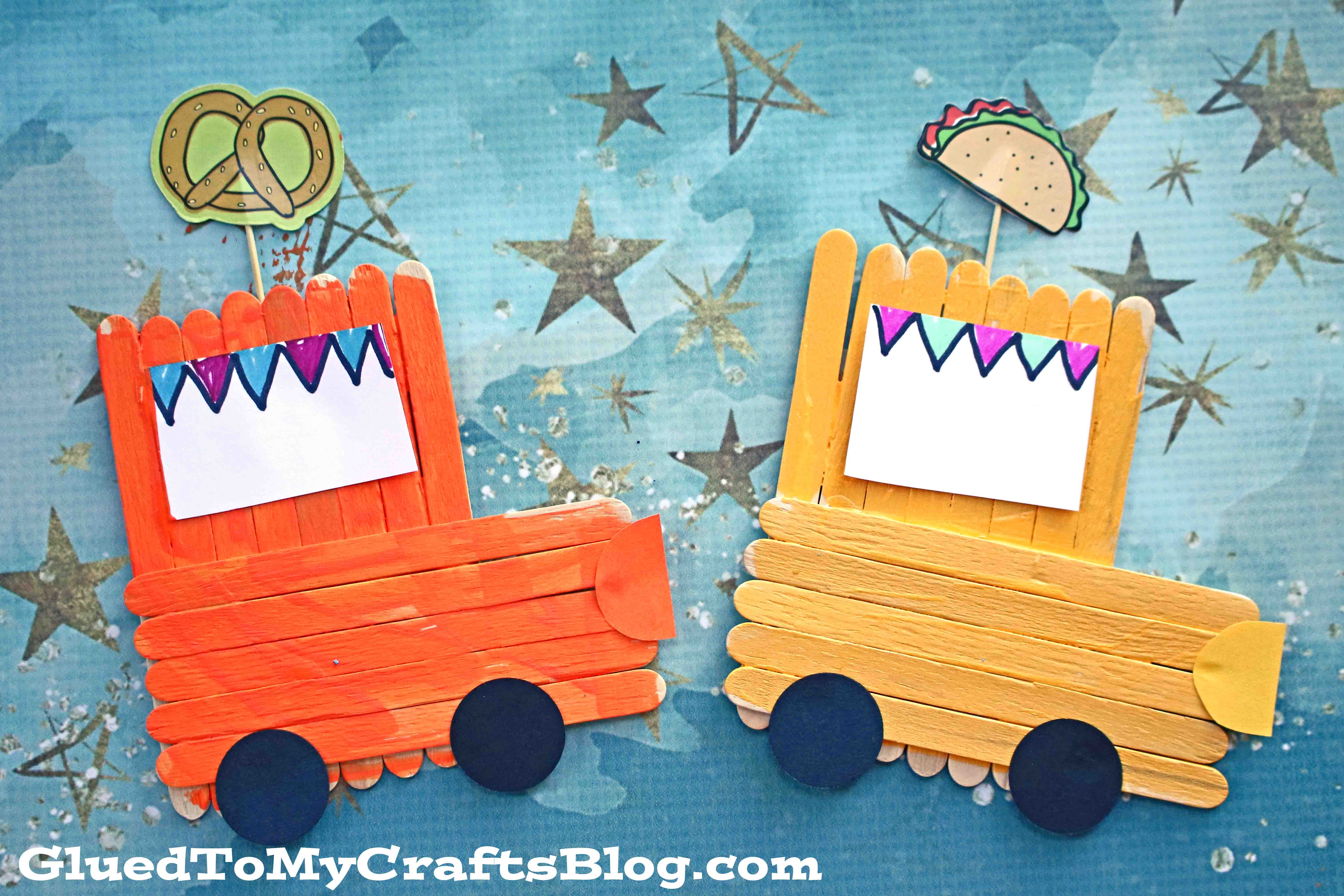Two colorful popsicle stick food trucks made from craft sticks and paper are displayed on a blue starry background—one orange with a pretzel, the other yellow with a taco. Both feature black wheels and festive paper banners.