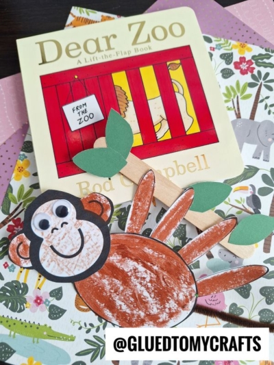 A children’s book titled “Dear Zoo” sits on floral and animal-themed paper. In front, there’s a handmade paper monkey craft made from a traced and painted handprint, with googly eyes and a smiling face. @GLUETOMYCRAFTS is written below.