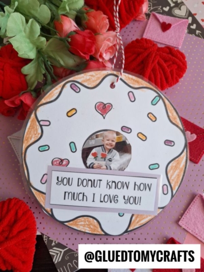 A decorated paper donut love frame with a child's photo in the center, surrounded by colored sprinkles, a heart cutout, and the message "You donut know how much I love you!" Red flowers are in the background.