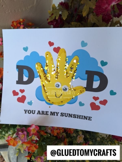 A handmade craft features a yellow handprint with googly eyes, glitter dots, and "dad you are my sunshine" surrounded by blue clouds and red hearts. Below, it says YOU ARE MY SUNSHINE, with flowers and leaves at the bottom.