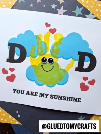 A handmade card features a blue cloud with a smiling yellow handprint sun, googly eyes, red hearts, large D letters, and the text "Dad you are my sunshine" on a starry background.