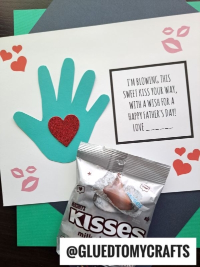 A handmade Fathers Day card features a teal paper handprint with a red glitter heart, a note saying I’m blowing kisses for daddy your way, with a wish for a Happy Father’s Day!, and a Hershey’s Kisses bag attached.