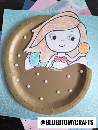 A paper plate clam craft made from a colored plate and a cutout mermaid with googly eyes. The plate is painted gold, decorated with pearls, and the mermaid’s arms and fins are brightly colored in.