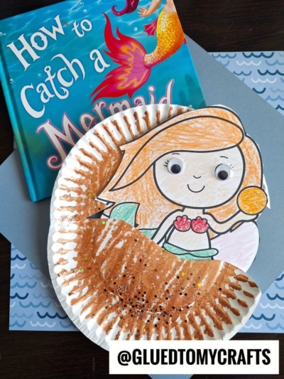 A children’s mermaid craft made with a colored mermaid cutout and a decorated paper plate clam, placed next to the book How to Catch a Mermaid on a table with wave-patterned paper. @GLUEDTOMYCRAFTS at the bottom.