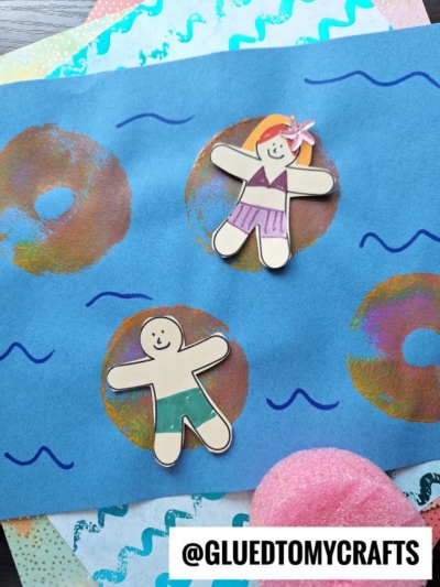 Two paper cutouts of smiling people in swimsuits are placed on blue paper with drawn waves, colorful rings, and pool noodle crafts, resembling water. The image includes the text @GLUEDTOMYCRAFTS at the bottom.